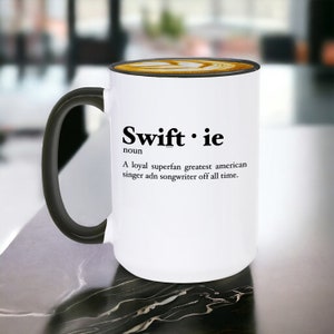 Pu&ograve; includere: Tazza in ceramica bianca con manico e bordo neri, con la scritta "Swiftie" e una definizione. La tazza contiene un latte con un disegno decorativo sulla parte superiore. La tazza &egrave; su una superficie di marmo.