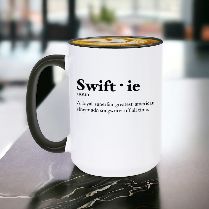 Pu&ograve; includere: Tazza in ceramica bianca con manico e bordo neri, con la scritta "Swiftie" e una definizione. La tazza contiene un latte con un disegno decorativo sulla parte superiore. La tazza &egrave; su una superficie di marmo.