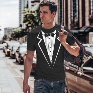 Pode incluir: Camiseta preta com um desenho de smoking branco. O desenho inclui uma gravata borboleta, uma camisa abotoada e um desenho de casaco. A pessoa usa jeans azuis e caminha na rua.