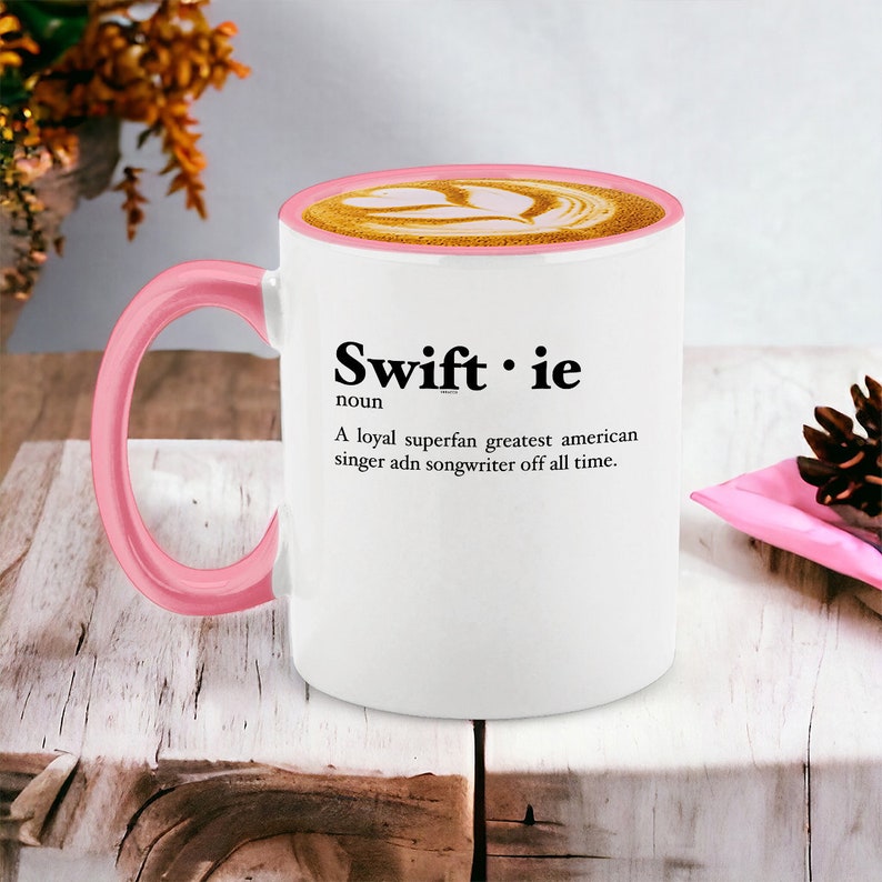 Pu&ograve; includere: Tazza da caff&egrave; bianca con manico e bordo rosa. La tazza presenta la parola "Swiftie" in grassetto nero, insieme a una definizione. La tazza &egrave; riempita con un latte con un design a cuore.
