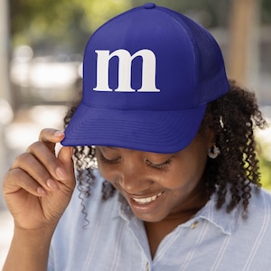 May include: A blue trucker hat with a white "m" on the front. The hat has a mesh back and a curved brim. The person in the image is wearing a light blue button-down shirt.