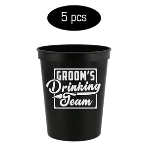 May include: Set of five black plastic cups with white text that reads "Groom's Drinking Team".