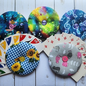 May include: Five round fabric pin cushions with decorative button centers. The cushions feature various patterns, including a blue and white gingham with sunflowers, a gray cat print, and galaxy and musical note designs. Playing cards are arranged around the cushions.