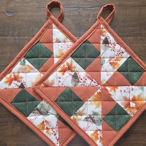 Op de afbeelding: Twee vierkante gewatteerde pannenlappen met een patchworkontwerp van effen roestoranje stof, donkergroene stof en een witte stof bedrukt met herfstbladeren in tinten oranje, geel en bruin. Elke pannenlap is voorzien van een stoffen ophanglus.