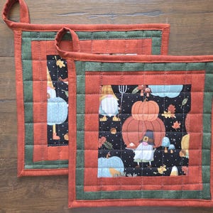 May include: Two square quilted pot holders featuring a central black fabric panel with a print of gnomes, pumpkins, and mushrooms. The borders are alternating bands of dark green and burnt orange fabric, with a fabric loop attached to one corner.