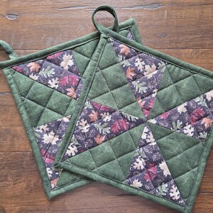 Könnte beinhalten: Zwei quadratische, gesteppte Topflappen mit einem Patchwork-Design aus einfarbig olivgrünem Stoff und einem dunklen Stoff mit einem Muster aus bunten Herbstblättern. Jeder Topflappen verfügt über eine Stoffschlaufe zum Aufhängen und Rautensteppung.