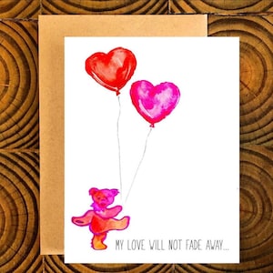 May include: A greeting card with a watercolor illustration of two heart-shaped balloons, one red and one pink, floating above a dancing bear. The text "MY LOVE WILL NOT FADE AWAY..." is printed at the bottom. The card is set against a brown envelope.