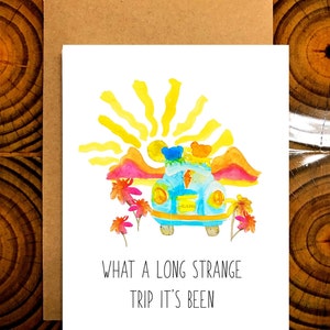 May include: Watercolor greeting card with a blue car, sun, and mountains. The car has a peace sign on the door and is decorated with flowers. The text on the card reads "WHAT A LONG STRANGE TRIP IT'S BEEN."