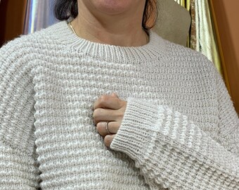 Könnte beinhalten: Ein cremefarbener Strickpullover mit einem strukturierten Muster. Der Pullover hat lange Ärmel und einen runden Ausschnitt.