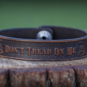May include: A brown leather bracelet with the words "Don't Tread On Me" embossed in copper. The bracelet has a silver metal clasp.