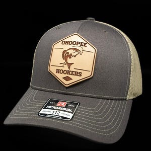 May include: A charcoal gray trucker hat with a beige mesh back and an adjustable snapback closure. A tan hexagonal leather patch is centered on the front, featuring a fish graphic and the text OHOOPEE HOOKERS. A black sticker is on the brim.