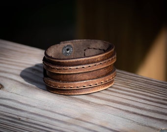 May include: A brown leather cuff bracelet with a single snap closure. The bracelet has two rows of stitching around the edges.