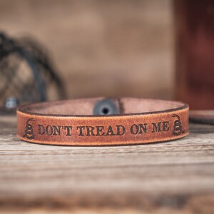 May include: A brown leather bracelet with the text "Don't Tread On Me" and a snake design.