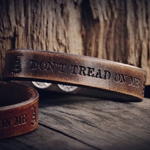 May include: Two brown leather bracelets with the phrase "DON'T TREAD ON ME" and a coiled snake symbol embossed on them. The bracelets are set against a rustic wooden background, showcasing a vintage aesthetic.
