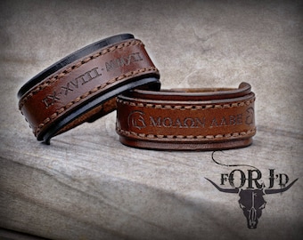 Puede incluir: Dos pulseras de cuero con costuras marrones. Una pulsera es negra con la inscripción "DXVIII.MMXXI" y la otra es marrón con la inscripción "MOAON AABE".