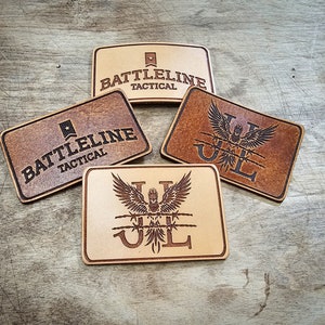 Könnte beinhalten: Vier rechteckige Lederaufnäher mit eingravierten Designs. Zwei Aufnäher tragen die Aufschrift "BATTLELINE TACTICAL" mit einem Sternemblem. Die anderen beiden zeigen einen stilisierten Adler mit den Buchstaben "JL". Die Aufnäher sind in verschiedenen Brauntönen gehalten.