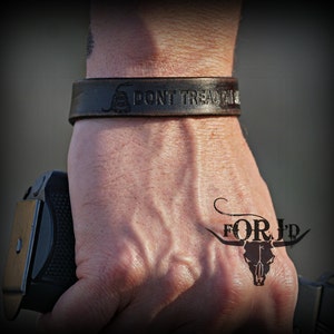 May include: A brown leather bracelet with the words "Don't Tread On Me" and a snake design. The bracelet is on a person's wrist, and there is a tattoo of a bull skull with the words "FOR JD" on the person's hand.