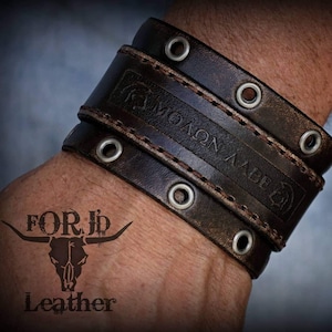 Puede incluir: Un brazalete de cuero marrón con múltiples filas de costuras y ojales plateados. El brazalete tiene el texto "FORJ" grabado.