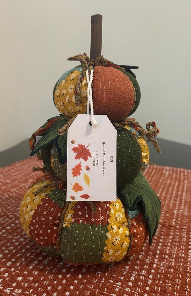 Pu&ograve; includere: Una decorazione a forma di zucca impilata e trapuntata con uno stelo di legno. La zucca &egrave; realizzata in tessuto di vari colori, tra cui arancione, giallo e verde. L'etichetta recita "Quilted Pumpkin Stacks" e le dimensioni sono 17.78cm x 17.78cm di larghezza e 29.21cm di altezza.