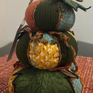 May include: A decorative, stacked pumpkin made of fabric. The pumpkin has three sections in shades of orange, green, yellow, and blue. Each section is adorned with stitched details and a twisted rope accent. A wooden stem and fabric leaves complete the fall-themed decoration.