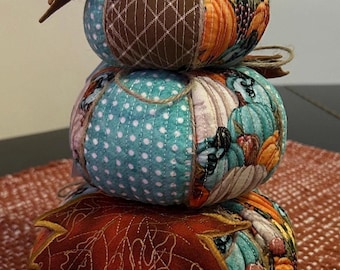 Può includere: Un centrotavola decorativo a forma di zucche impilate. Le tre zucche di stoffa sono decorate con motivi a tema autunnale, tra cui pois, zucche e foglie. Ogni zucca è fissata con spago e sormontata da uno stelo marrone e accenti di foglie.