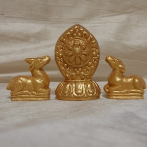 May include: A set of three gold-colored figurines consisting of two identical deer statues in a reclining position and one central upright emblem featuring a circular wheel design with intricate petal-like carvings on a pedestal base.