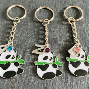 May include: Three silver keychains with panda bear charms. Each panda is holding a green bamboo shoot and has a letter charm with a jewel on top. The letters are A, Z, and D. The jewels are blue, purple, and pink.