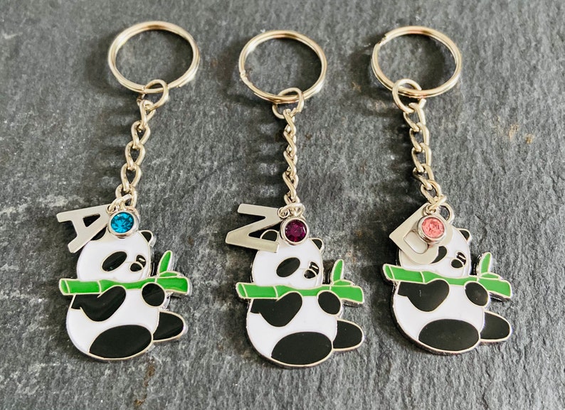May include: Three silver keychains with panda bear charms. Each panda is holding a green bamboo shoot and has a letter charm with a jewel on top. The letters are A, Z, and D. The jewels are blue, purple, and pink.