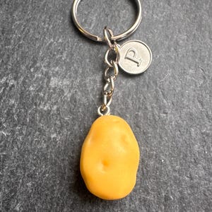 May include: A novelty keyring featuring a small, yellow potato charm. The charm is attached to a silver chain and a round tag with the letter "P". The keyring has a silver ring.