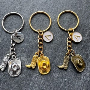 May include: Three keyrings with cowboy-themed charms. One keyring has a silver ring, chain, and charms, including a boot, a cowboy hat, and a circular charm with the letter "A". The other two keyrings have gold rings, chains, and charms.