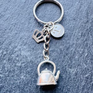 May include: Silver teapot keyring with a crown and a charm with the letter T. The keyring is attached to a silver key ring.