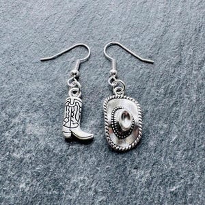 May include: Silver-coloured cowboy boot and cowboy hat drop earrings. The earrings feature detailed designs, with the boot showcasing intricate patterns and the hat having a rope-like border. The earrings are suspended from simple ear wires.