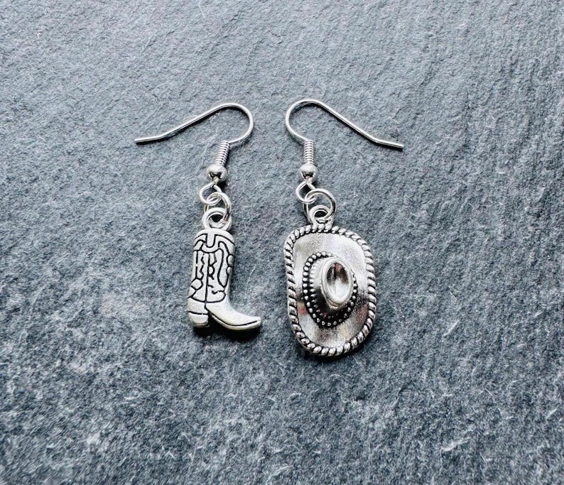 May include: Silver-coloured cowboy boot and cowboy hat drop earrings. The earrings feature detailed designs, with the boot showcasing intricate patterns and the hat having a rope-like border. The earrings are suspended from simple ear wires.