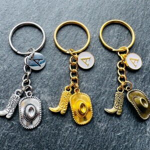 May include: Three keyrings with cowboy-themed charms. Each keyring has a ring, a charm with a letter, a boot charm, and a cowboy hat charm. The keyrings are silver and gold in colour.