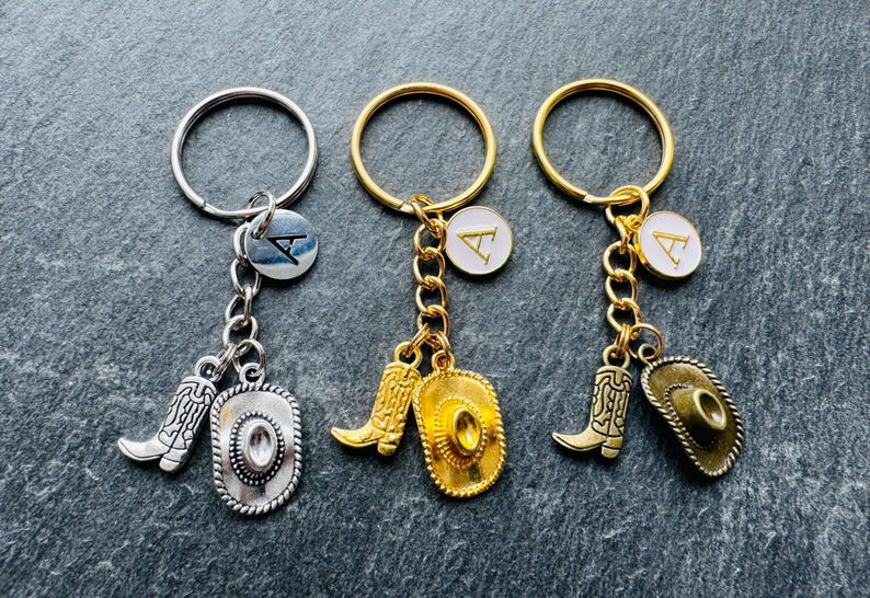 May include: Three keyrings with cowboy-themed charms. Each keyring has a ring, a charm with a letter, a boot charm, and a cowboy hat charm. The keyrings are silver and gold in colour.