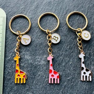 May include: Three gold keychains with giraffe charms. The charms are yellow, pink, and white with black spots. Each keychain has a white circle charm with a gold letter, A, B, and C.