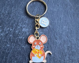 May include: A gold-coloured keyring featuring a cartoon mouse holding a piece of cheese. The mouse is brown with pink ears and a blue belly. A small, round charm with the letter "R" is also attached.