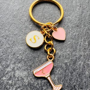 May include: Gold-coloured keyring with a round ring and multiple charms. Charms include a white circle with a gold "S", a pink heart, and a pink cocktail glass. The cocktail glass charm has white accents.