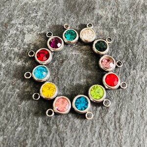 May include: A set of 12 silver-toned charms with colorful crystal accents. Each charm features a small loop for attaching to jewelry or other crafts.