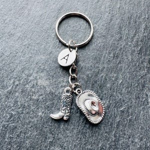 May include: Silver-coloured keyring with charms. The charms include a cowboy boot, a cowboy hat, and a circular charm with the letter "A". The keyring is attached to a silver ring. The charms have a Western theme.