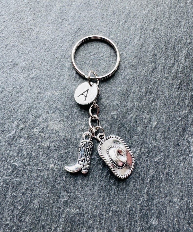 May include: Silver-coloured keyring with charms. The charms include a cowboy boot, a cowboy hat, and a circular charm with the letter "A". The keyring is attached to a silver ring. The charms have a Western theme.