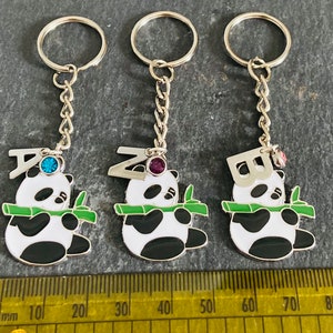 May include: Three silver keychains with panda bear charms. Each panda is holding a green bamboo stalk and has a letter charm on top. The letters are A, Z, and B. Each charm has a small jewel on top: blue, purple, and pink.