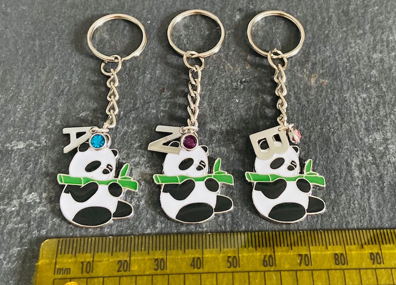 May include: Three silver keychains with panda bear charms. Each panda is holding a green bamboo stalk and has a letter charm on top. The letters are A, Z, and B. Each charm has a small jewel on top: blue, purple, and pink.
