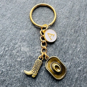 May include: Gold-coloured keyring with a cowboy theme. The keyring features a gold ring, a charm with the letter "A" on a white background, a cowboy boot charm, and a cowboy hat charm.