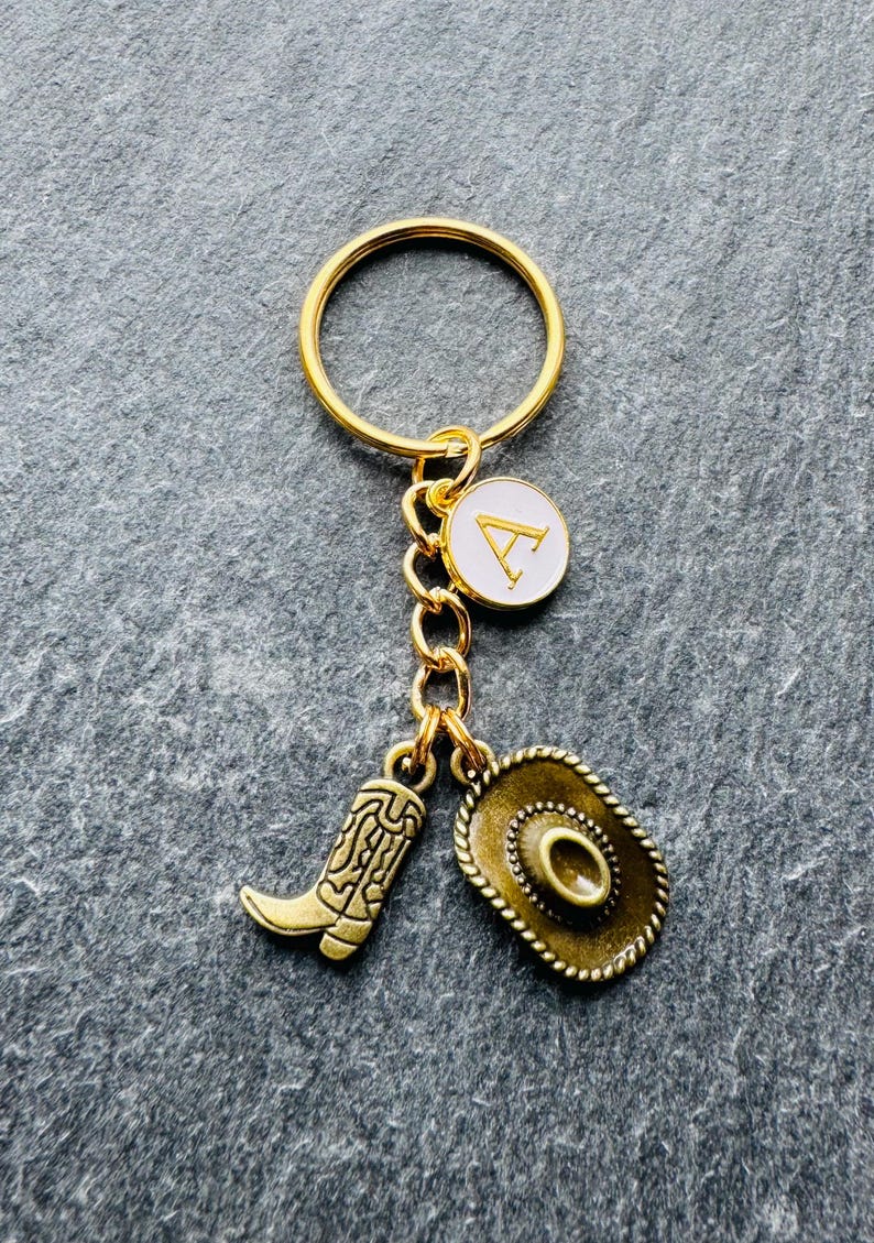 May include: Gold-coloured keyring with a cowboy theme. The keyring features a gold ring, a charm with the letter "A" on a white background, a cowboy boot charm, and a cowboy hat charm.