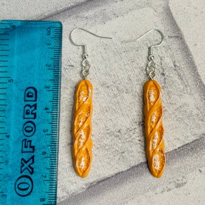 May include: A pair of dangle earrings featuring a realistic-looking baguette bread charm. The charms are a light brown color with a slightly darker brown swirl pattern. The earrings are made with silver-toned hooks.