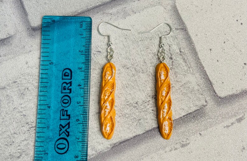 May include: A pair of dangle earrings featuring a realistic-looking baguette bread charm. The charms are a light brown color with a slightly darker brown swirl pattern. The earrings are made with silver-toned hooks.