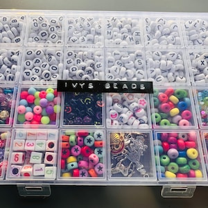 May include: A clear plastic storage box filled with various colourful beads and letter beads. The box has 18 compartments and is labelled "IVYS BEADS".