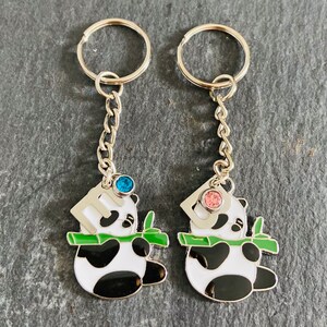 May include: Two silver keychains with panda bear charms. The panda bears are holding bamboo stalks and have blue and pink rhinestones on their heads.
