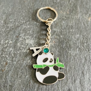 May include: A silver keychain with a panda bear charm holding a piece of bamboo. The panda has a black and white face with a green gemstone on its head. The keychain has a silver letter "A" charm.